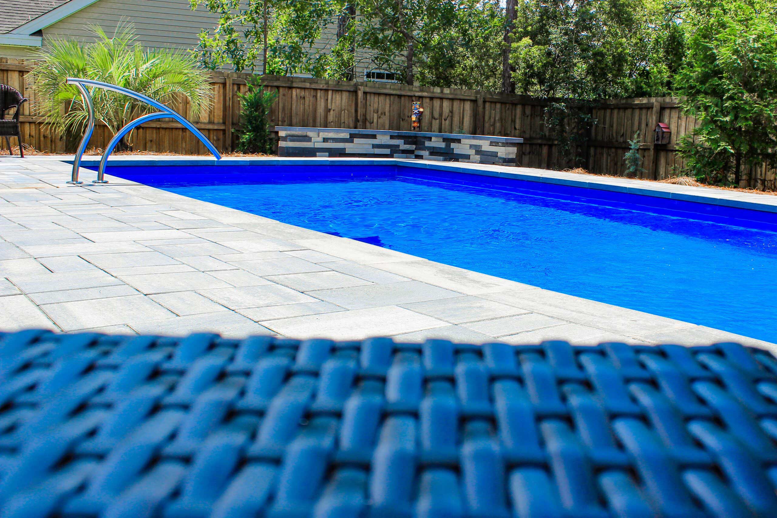 A blue pool in a fenced backyard taken from the POV of a pool lounge chair